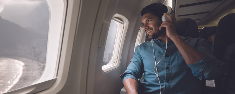 A man wearing headphones smiles while looking out the window of an airplane during a flight.
