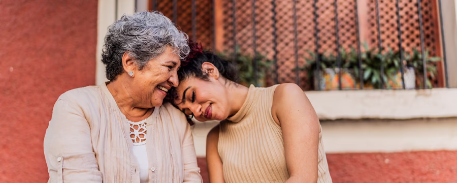 Two women, an older woman and younger woman, sitting together against a terracotta-colored wall. They are holding hands, leaning close with their heads together in an intimate, caring moment. Both are smiling warmly. Soft green foliage is visible in the background.