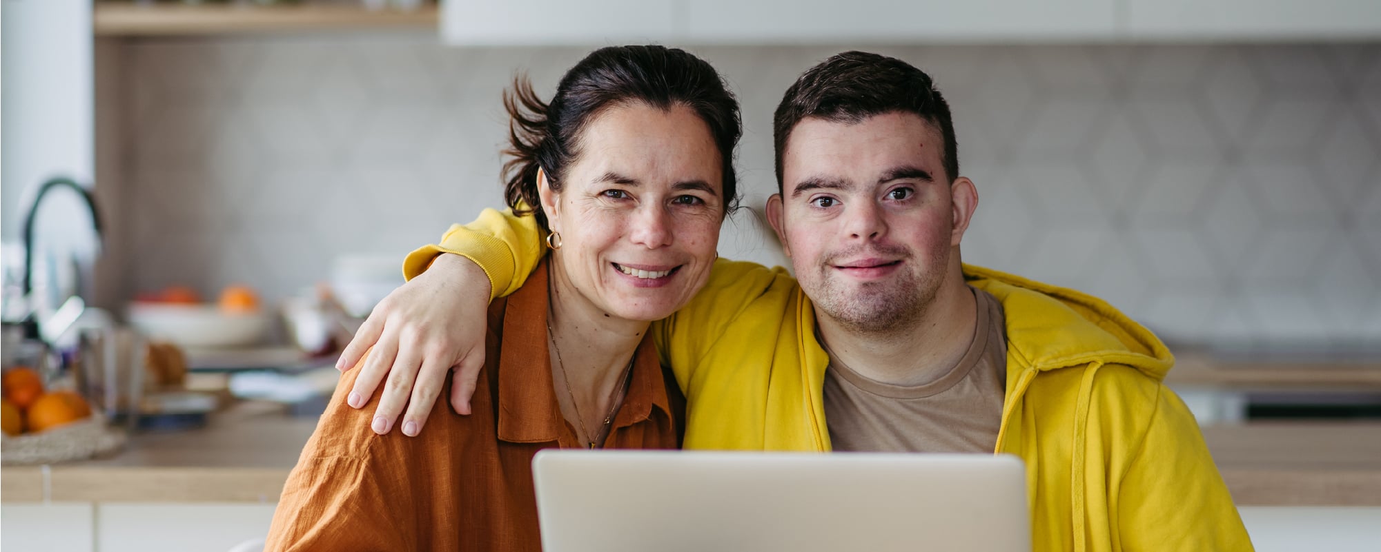 Woman and young man sitting close together at a kitchen table, smiling at the camera behind an open laptop.