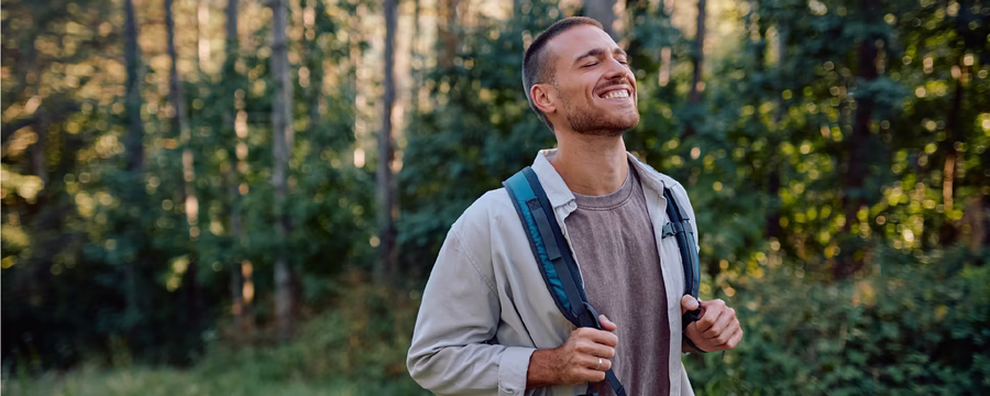 Smiling man wearing a backpack enjoys a peaceful hike along a sunlit forest trail.