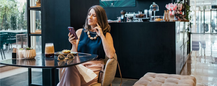 Woman sitting at a café table, smiling as she looks at her smartphone with an iced coffee beside her.