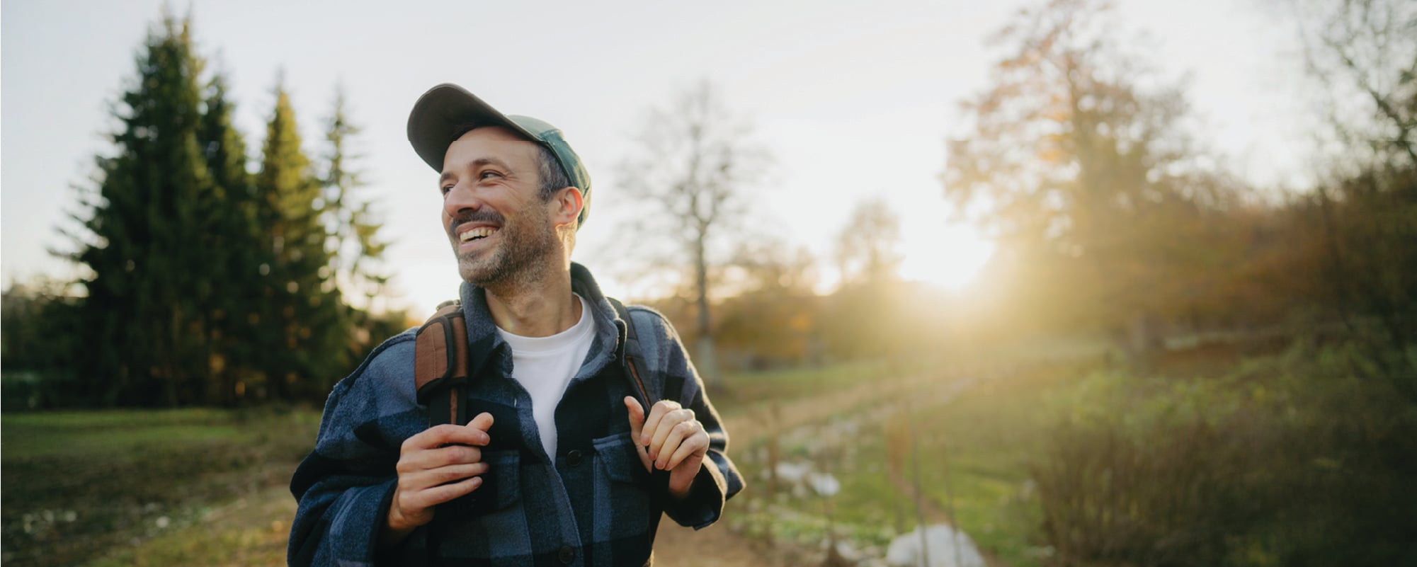 A smiling person wearing a baseball cap and backpack walks along a sunlit nature trail, looking off into the distance. Trees, greenery, and warm golden-hour light create an inviting outdoor setting.