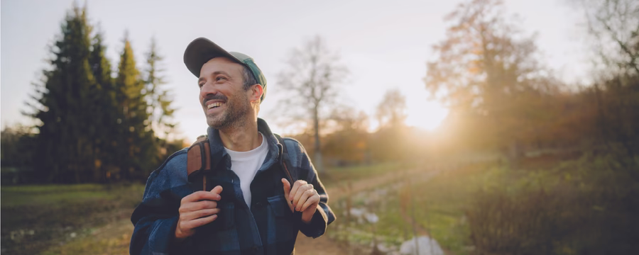 A smiling person wearing a baseball cap and backpack walks along a sunlit nature trail, looking off into the distance. Trees, greenery, and warm golden-hour light create an inviting outdoor setting.