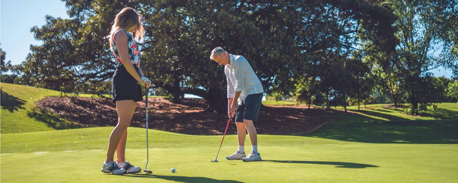 Two people stand on a golf green preparing to putt on a sunny day, with large trees and rolling grass in the background.