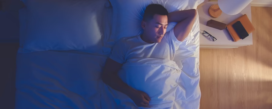 A man lying in bed under white sheets, resting on his back with one arm behind his head, illuminated by soft blue nighttime lighting, with a bedside table and lamp nearby.