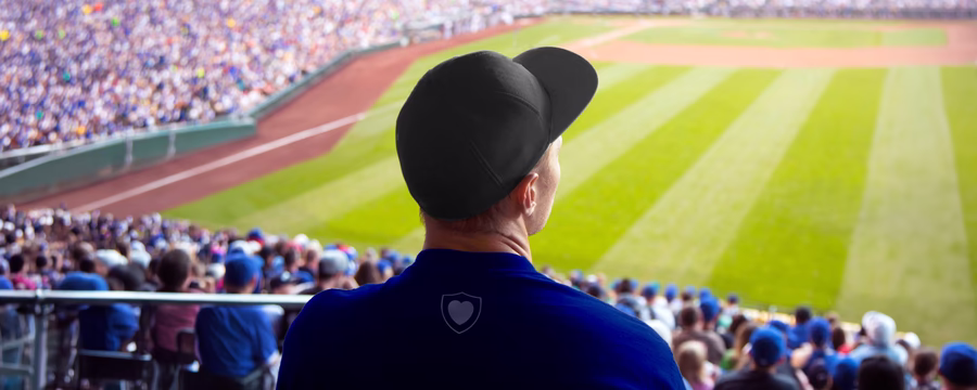 A man wearing a baseball cap sits in the stands, watching a game on a crowded baseball field.
