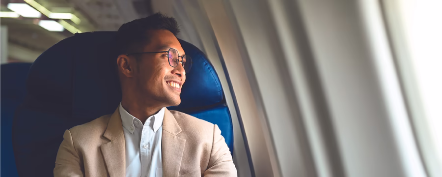 A smiling man wearing glasses and a beige blazer sits by an airplane window, looking outside while resting his hands together on the armrest.
