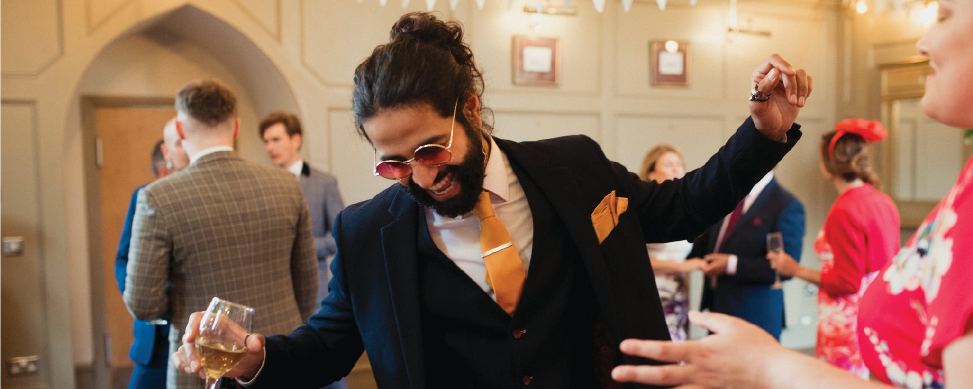 A smiling man in a dark suit with a bright orange tie and pocket square dances while holding a glass of white wine at a lively indoor celebration. Other well-dressed guests mingle in the background, creating a festive and energetic atmosphere.
