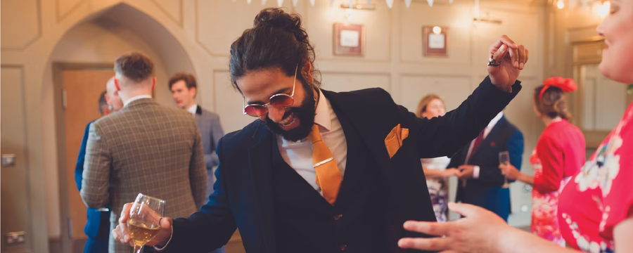 A smiling man in a dark suit with a bright orange tie and pocket square dances while holding a glass of white wine at a lively indoor celebration. Other well-dressed guests mingle in the background, creating a festive and energetic atmosphere.