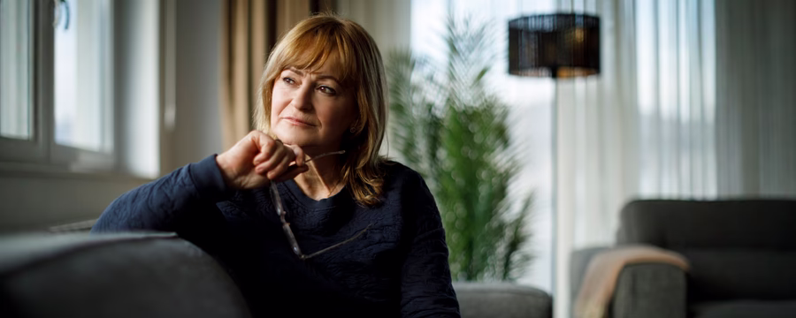 Older woman sitting on a sofa by a window, holding her eyeglasses and looking thoughtfully to the side.