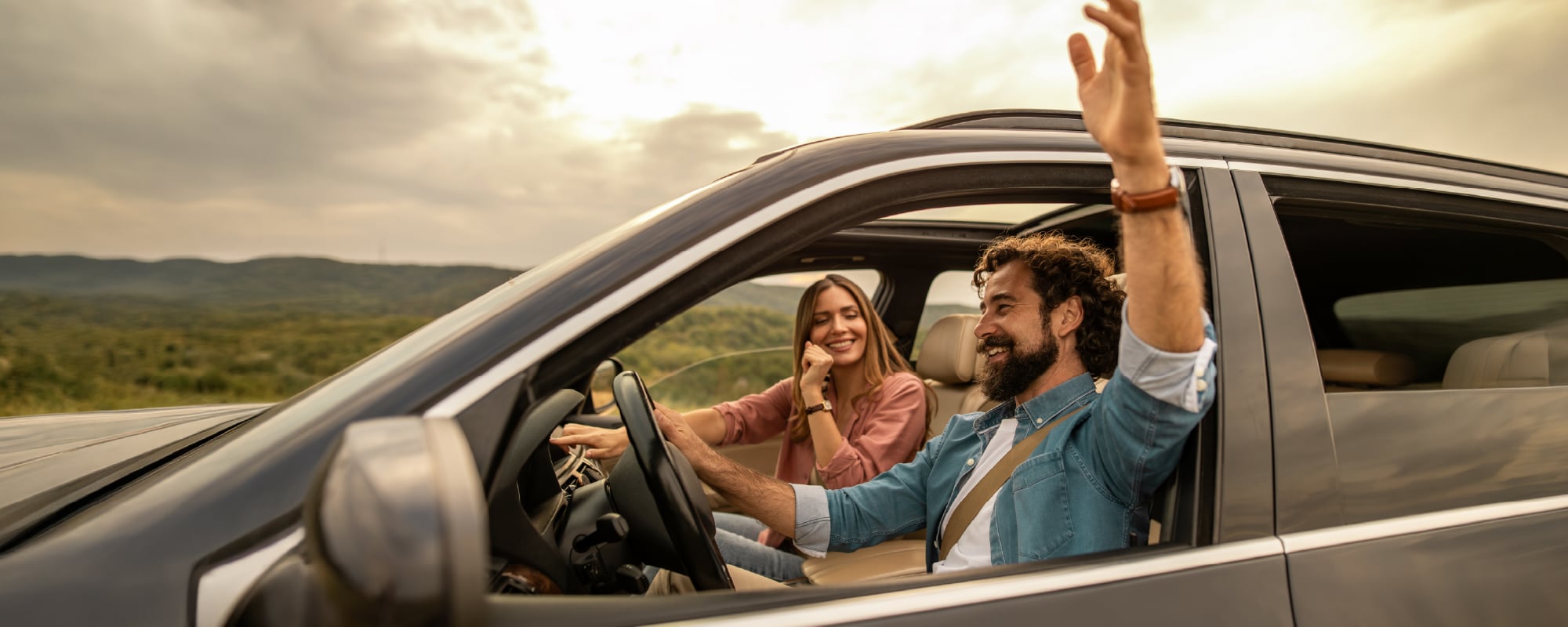 A smiling couple sits in a modern SUV parked at a scenic overlook during golden hour. The driver, wearing a seat belt, waves enthusiastically out the open window while the passenger smiles beside him. Rolling green hills and a warm sunset sky create a relaxed road trip atmosphere.