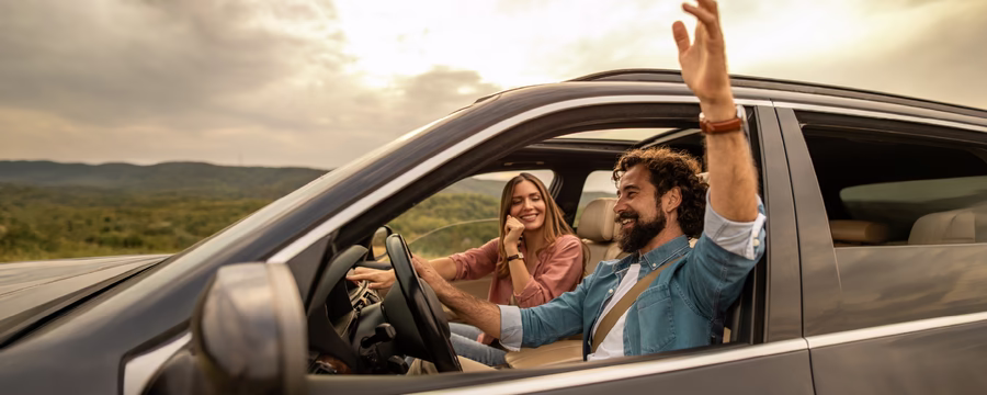 A smiling couple sits in a modern SUV parked at a scenic overlook during golden hour. The driver, wearing a seat belt, waves enthusiastically out the open window while the passenger smiles beside him. Rolling green hills and a warm sunset sky create a relaxed road trip atmosphere.