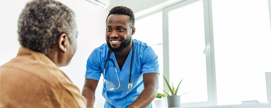 Smiling healthcare worker in blue scrubs and a stethoscope talking with an older man in a bright clinical setting.