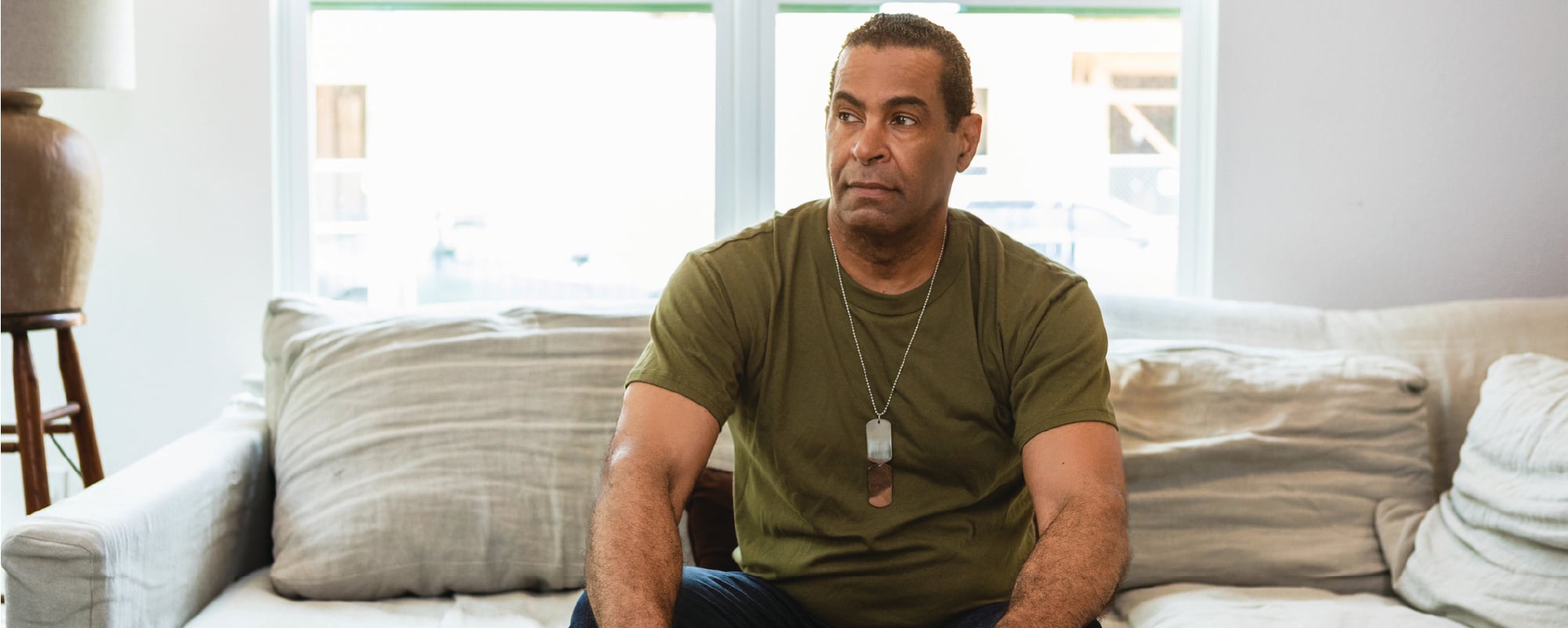 A man wearing a green T-shirt and military-style dog tags sits on a light-colored couch in a bright living room, looking thoughtfully off to the side with his hands resting on his knees.