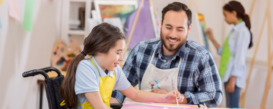 A young wheelchair user paints on a canvas during an art therapy session while a smiling instructor offers guidance, with art supplies on the table and another participant painting in the background.