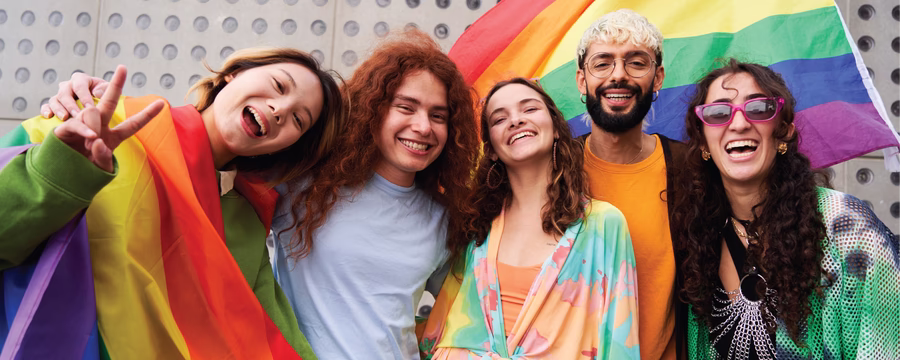 Five smiling people pose together outdoors in front of a large rainbow pride flag. One person is wrapped in a rainbow flag, and the group wears colorful clothing while looking at the camera and celebrating.