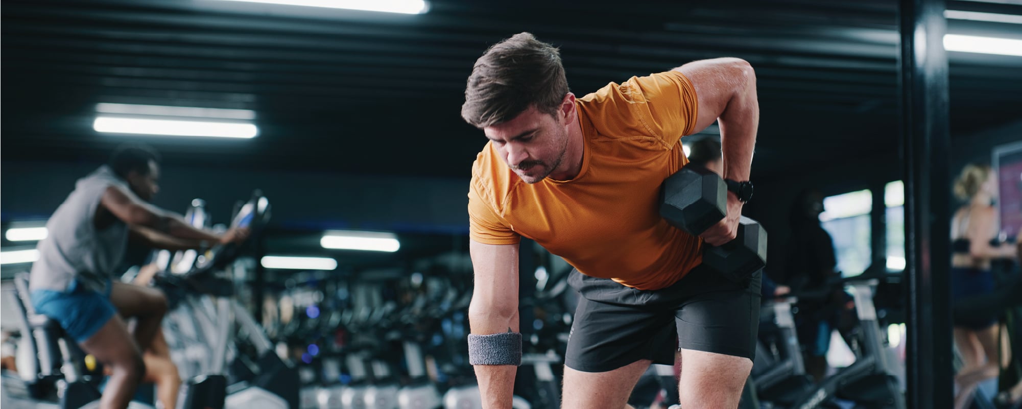A man performs a one-arm dumbbell row on a weight bench in a gym, wearing an orange athletic shirt and black shorts. Cardio machines and workout equipment are visible in the softly blurred background, emphasizing a focused strength training session.