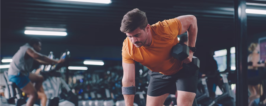 A man performs a one-arm dumbbell row on a weight bench in a gym, wearing an orange athletic shirt and black shorts. Cardio machines and workout equipment are visible in the softly blurred background, emphasizing a focused strength training session.