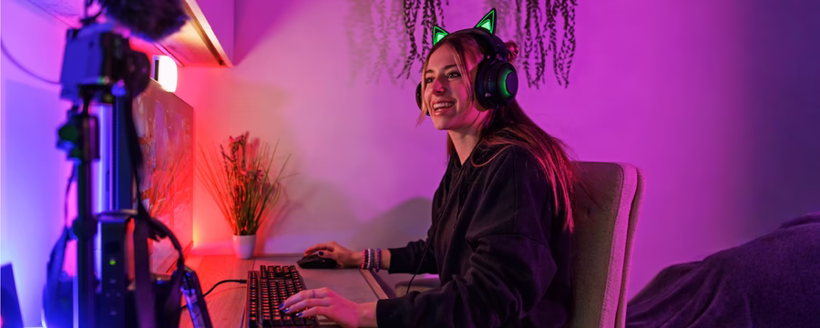 Alt text: Young woman wearing cat-ear gaming headphones smiles at a computer while using a keyboard and mouse in a colorful purple and red-lit gaming setup.