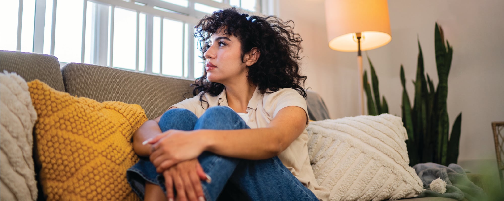 A woman with curly dark hair sits curled up on a couch, looking toward a bright window, with colorful pillows and a lit floor lamp around her.