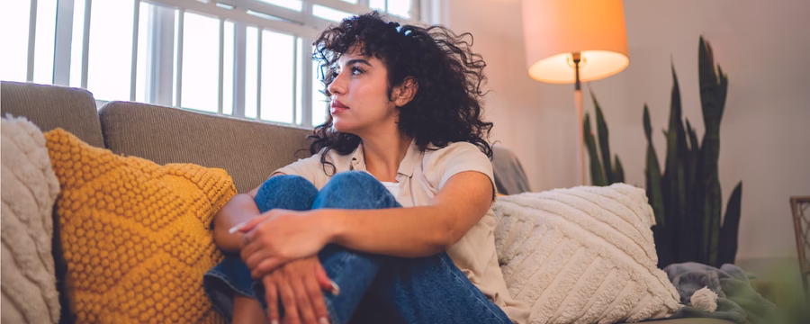 A woman with curly dark hair sits curled up on a couch, looking toward a bright window, with colorful pillows and a lit floor lamp around her.