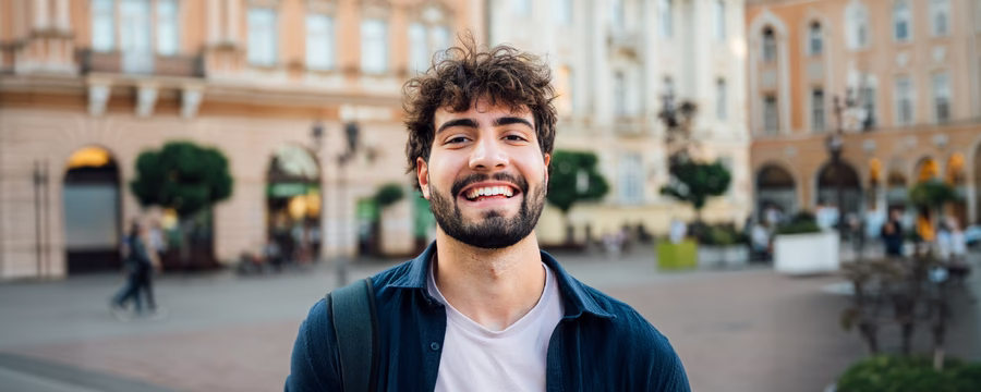Smiling young adult with curly dark hair and a beard stands in a European-style city square, wearing a white T-shirt under a navy overshirt and a backpack, with historic buildings softly blurred in the background.