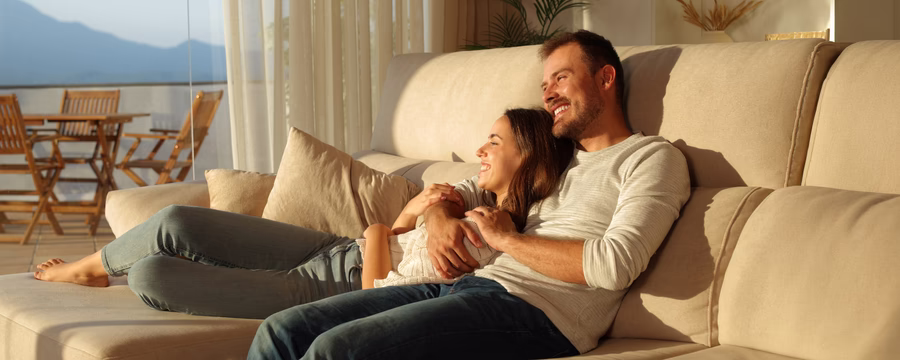 A smiling couple relaxes on a beige couch in warm sunlight, with the woman leaning against the man as he wraps his arm around her, both looking content and comfortable in a cozy living room setting.