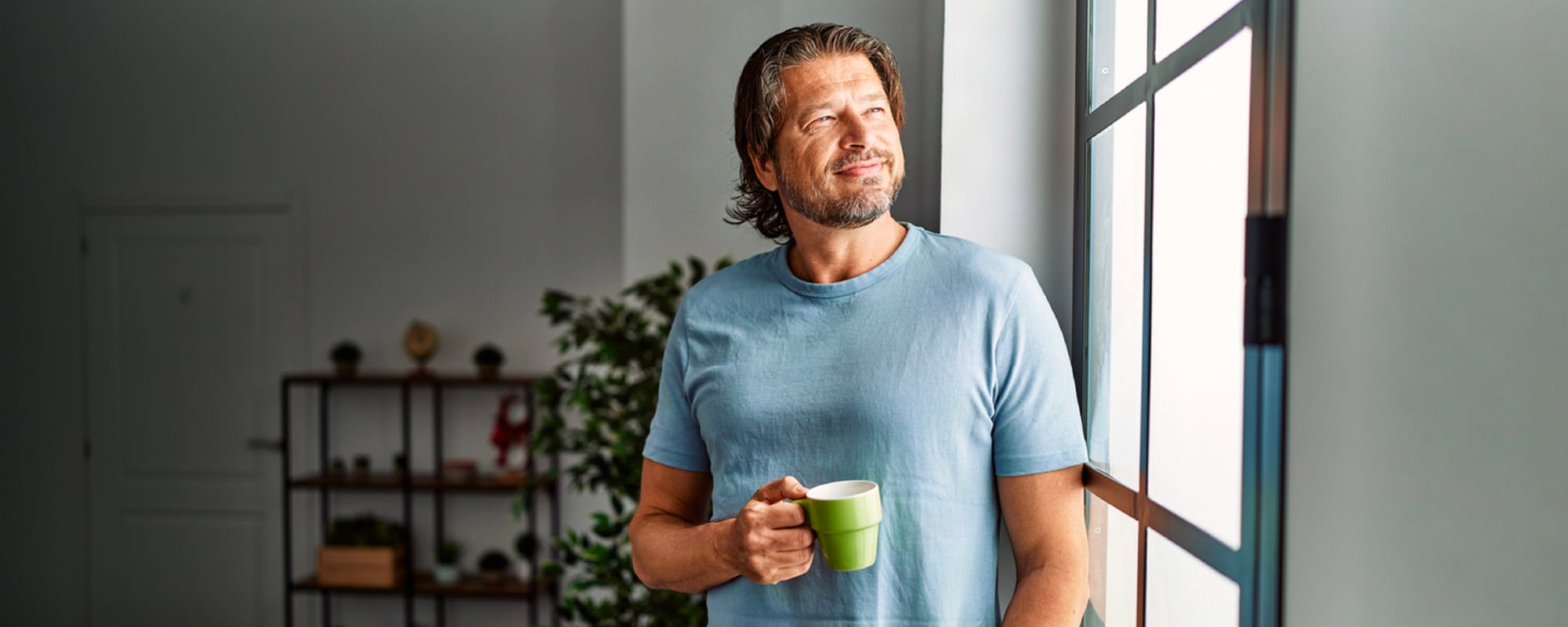 Smiling man in a light blue T-shirt holding a green mug while standing beside a sunlit window.