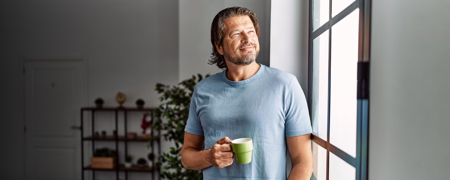 Smiling man in a light blue T-shirt holding a green mug while standing beside a sunlit window.