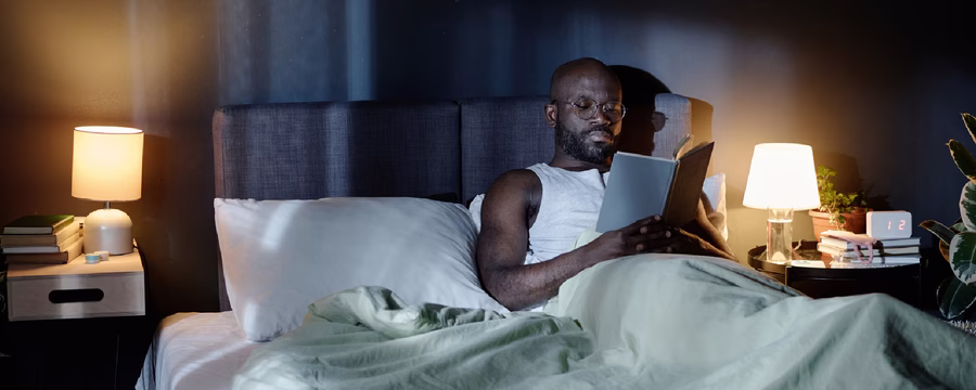 A man wearing glasses sits up in bed at night, reading a book beside a glowing bedside lamp.
