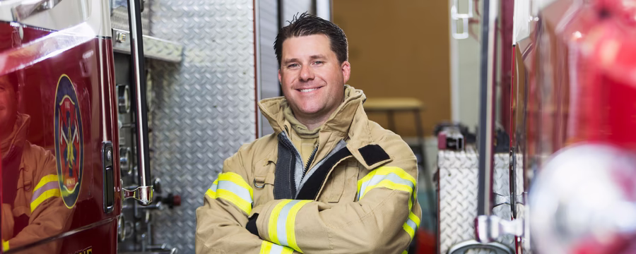 A smiling firefighter in tan turnout gear stands with arms crossed between fire trucks at a fire station.