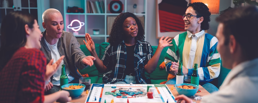 Five adults gathered around a table, laughing and socializing while playing a colorful board game in a cozy living room.