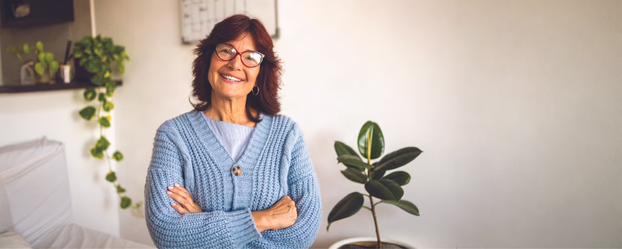 Smiling older woman wearing glasses and a light blue cardigan, standing with her arms crossed in a bright room with houseplants.