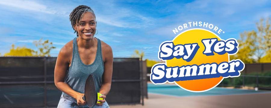 A smiling woman holds a pickleball paddle and ball on an outdoor court under a blue sky. A bright "NorthShore Say Yes Summer" logo appears beside her, promoting an active, confident lifestyle.
