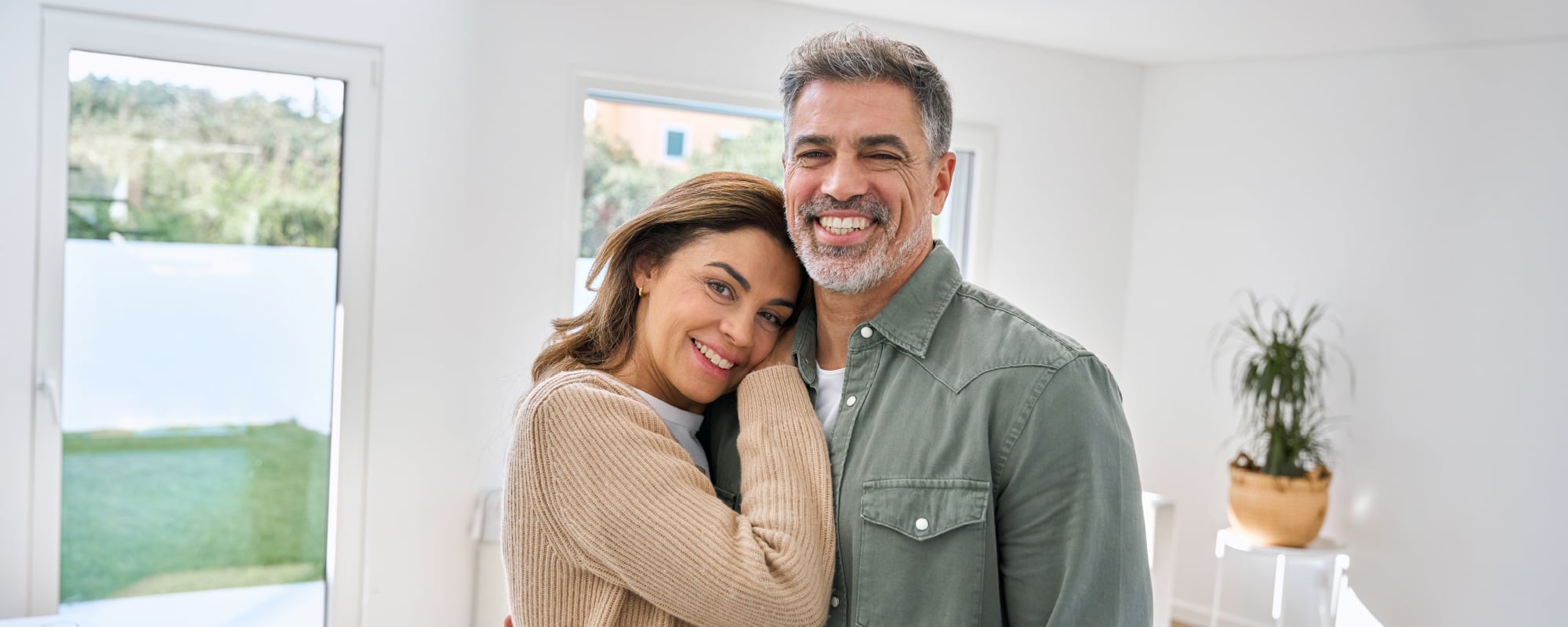 Smiling middle-aged couple embraces in a bright, sunlit home, with the woman resting her head on the man's shoulder as they look at the camera.