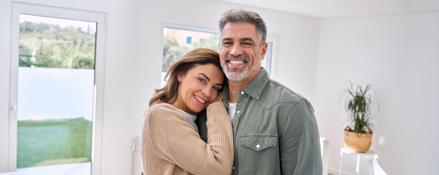 Smiling middle-aged couple embraces in a bright, sunlit home, with the woman resting her head on the man's shoulder as they look at the camera.
