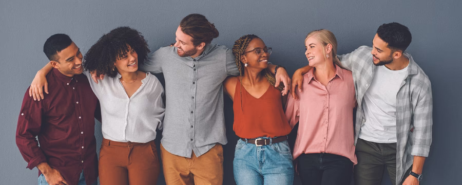 Six smiling adults of diverse backgrounds stand side by side with their arms around each other's shoulders, sharing a friendly moment against a plain blue wall. They wear casual clothing and look at one another, conveying connection, support, and inclusivity.