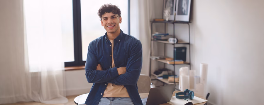 A smiling man in a blue shirt stands with his arms crossed in a bright room, with a desk, laptop, and headphones nearby.