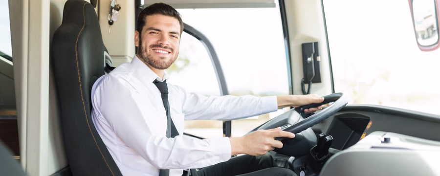 Smiling bus driver wearing a white dress shirt and black tie, seated behind the steering wheel inside a modern bus, looking toward the camera. The driver's seat, dashboard, and large front windows are visible, with bright daylight illuminating the interior.