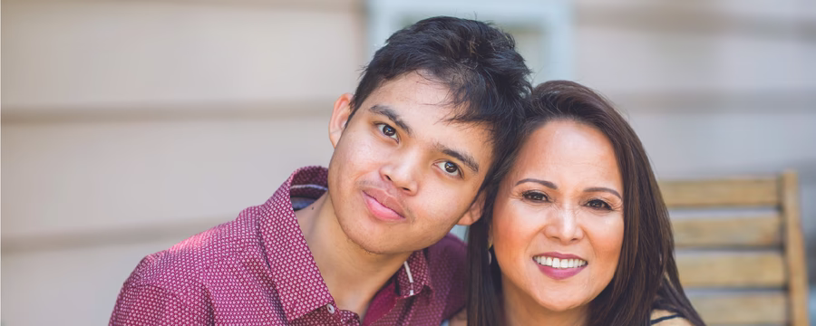 A smiling woman and a teenage boy sit close together outdoors, leaning their heads together and looking at the camera, with a wooden bench and house siding in the background.
