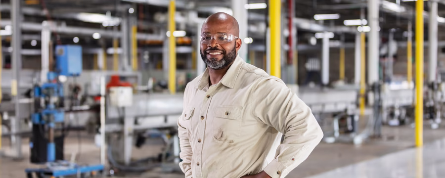 A smiling manufacturing worker wearing safety glasses stands with hands on hips inside a factory, with industrial equipment and production machinery blurred in the background.