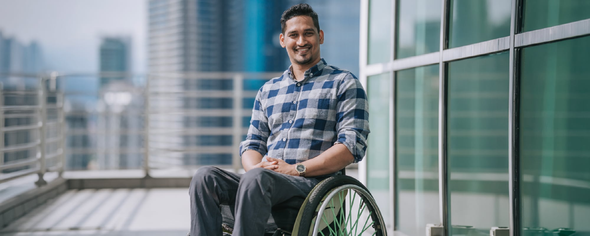 Smiling person seated in a wheelchair on an outdoor terrace in front of modern office buildings, wearing a blue plaid shirt and looking at the camera.