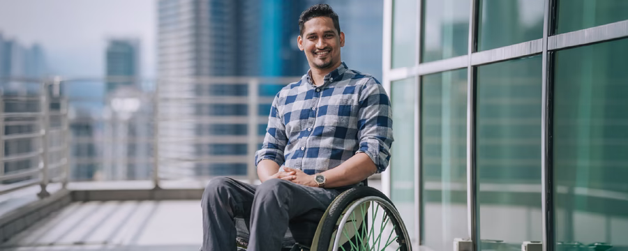 Smiling person seated in a wheelchair on an outdoor terrace in front of modern office buildings, wearing a blue plaid shirt and looking at the camera.