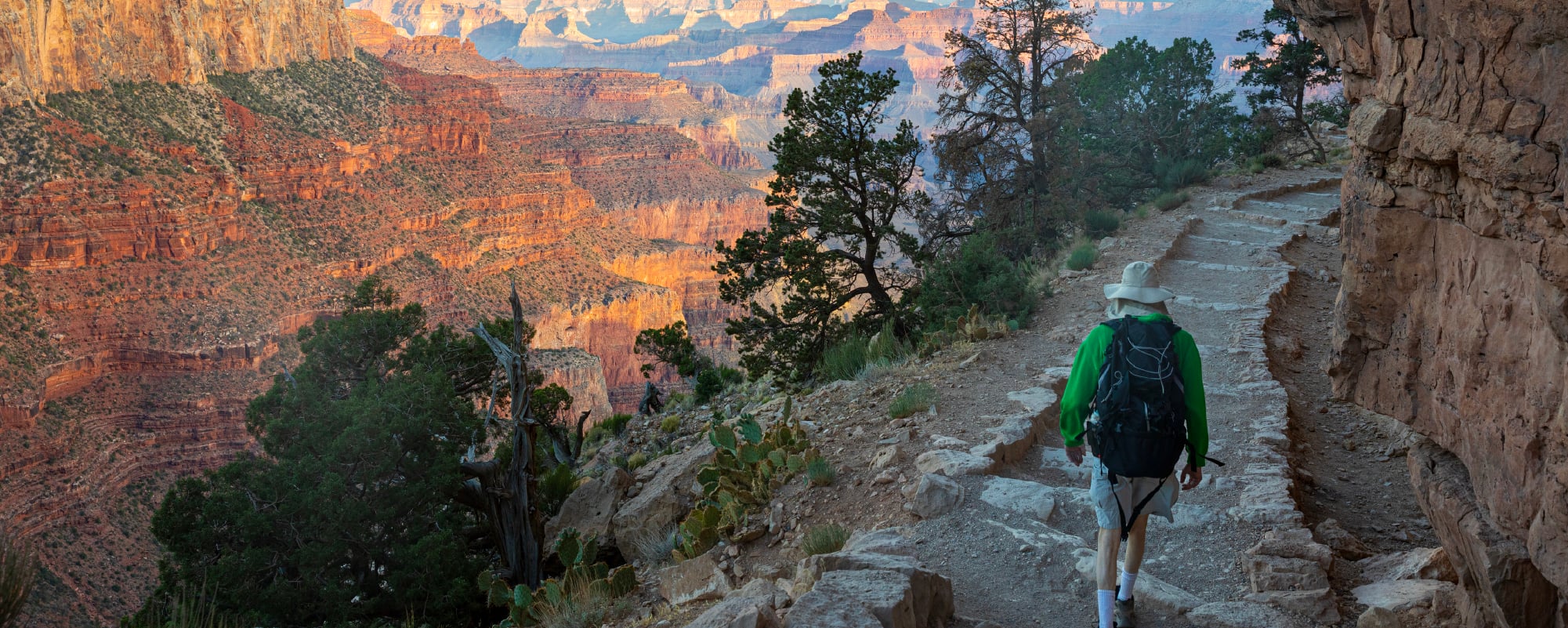 Hiker with a backpack descends a rocky cliffside trail overlooking the vast red-orange canyons of the Grand Canyon.
