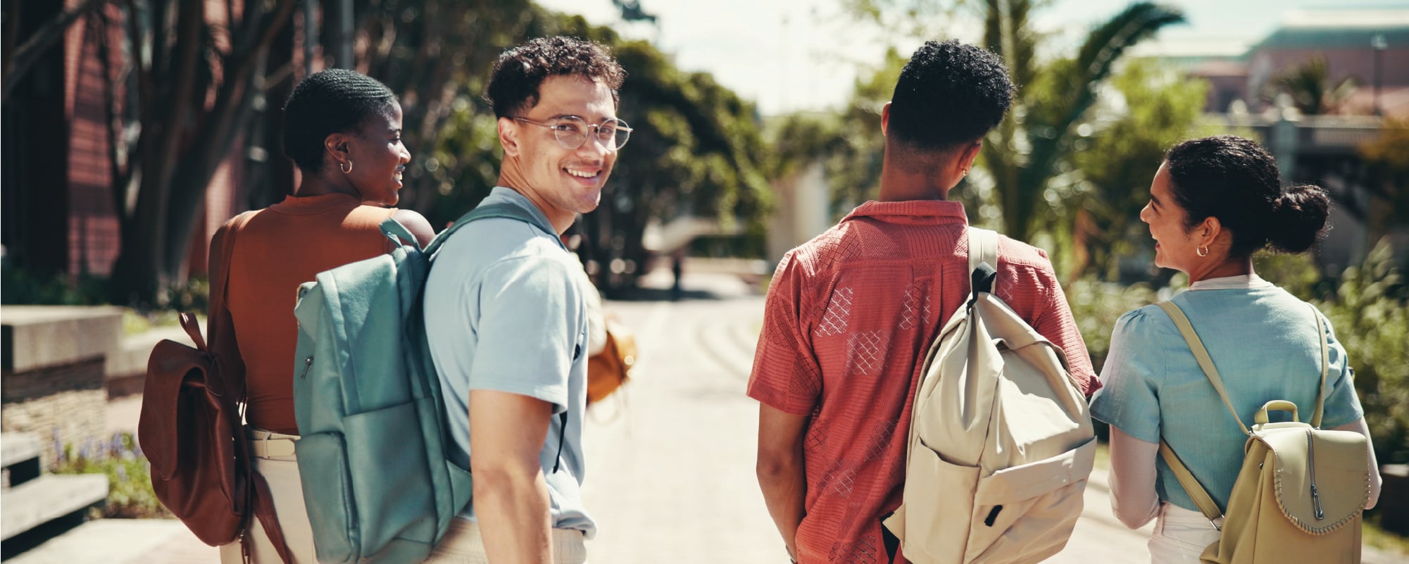 Four friends with backpacks walk along a sunny, palm-lined path while one turns back and smiles at the camera.