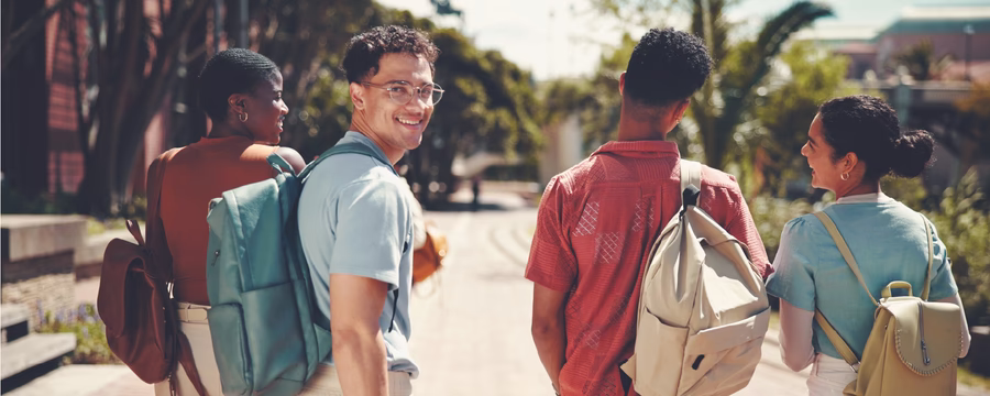 Four friends with backpacks walk along a sunny, palm-lined path while one turns back and smiles at the camera.