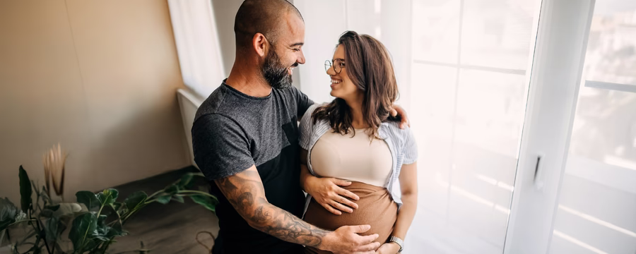 A smiling pregnant woman and her partner stand together by a sunlit window in their home, gazing at each other while both gently cradle her baby bump. The bright, softly lit room creates a warm, intimate atmosphere.