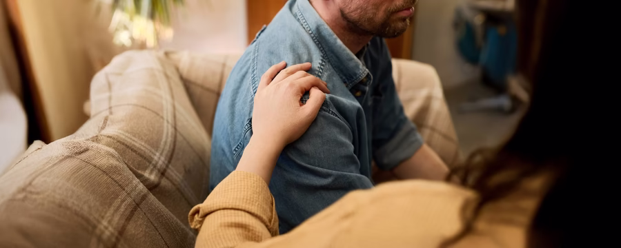 Supportive hand on a man's shoulder while sitting on a couch, symbolizing empathy, understanding, and compassionate care for incontinence challenges.