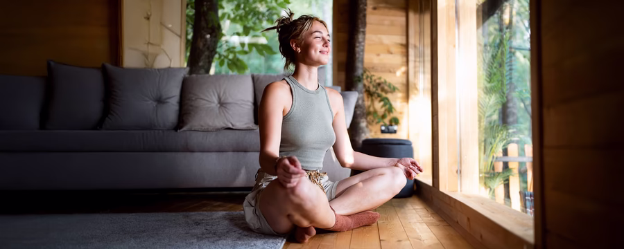 Woman meditating peacefully on floor in cozy sunlit living room with nature view