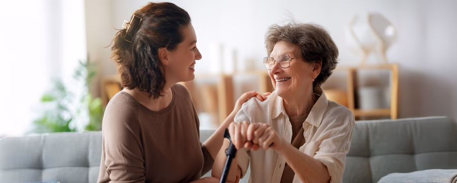 Young woman smiling and supporting an elderly woman using a cane, showing caregiver encouragement and support.